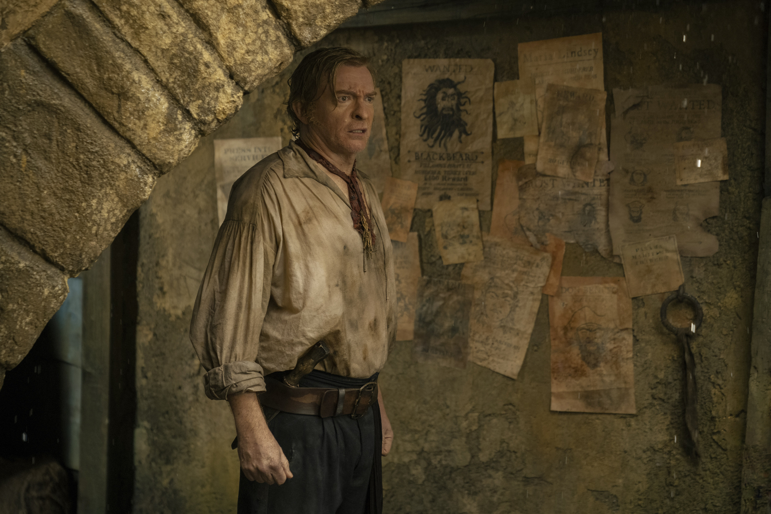 Rhys Darby as Stede Bonnet in Our Flag Means Death Season 2 looking the least fancy he’s ever been in a ragged stained formerly white blouse and black trousers, standing in what looks like a cellar (HBO Max)
