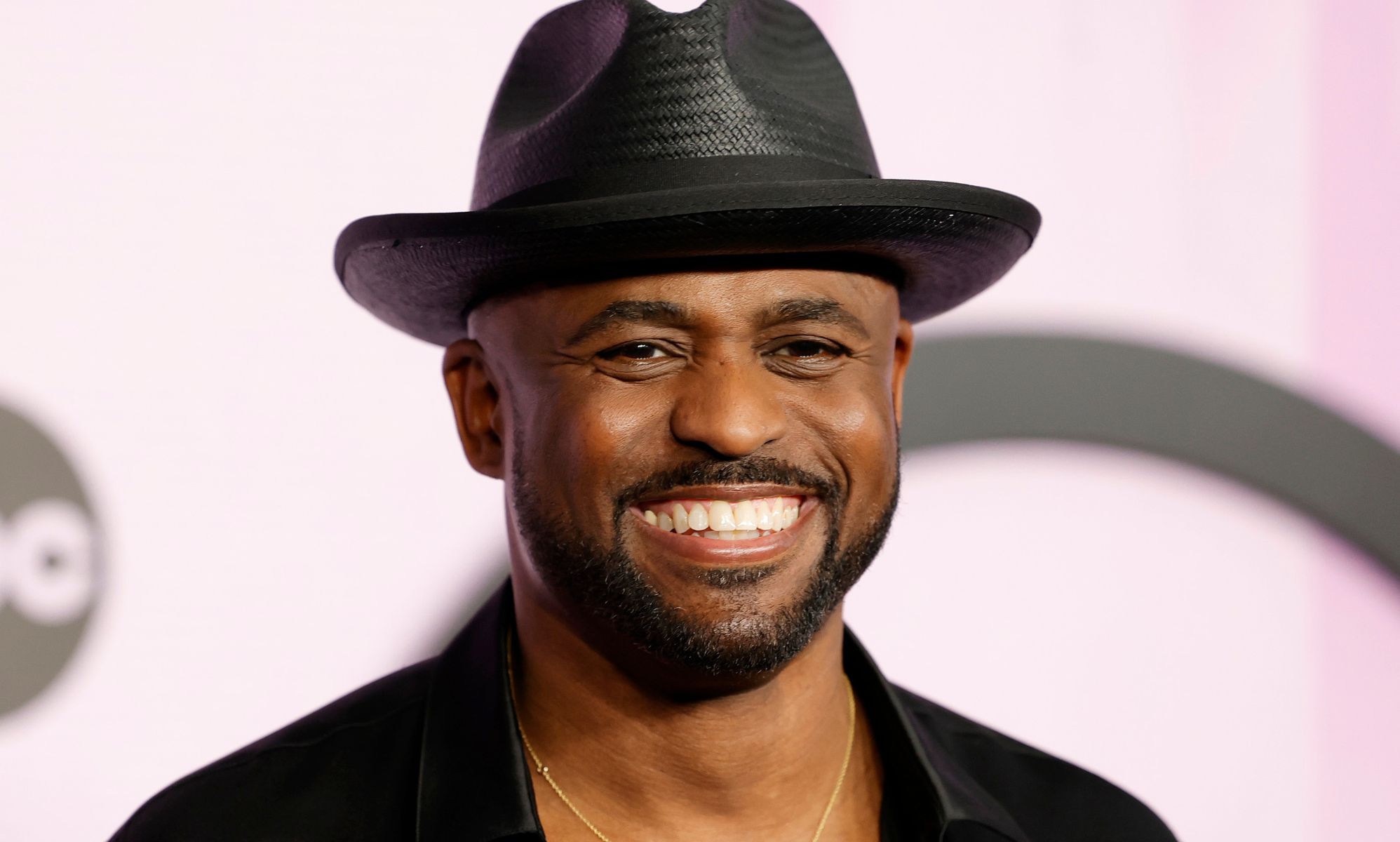 Wayne Brady smiles while wearing a black hat and black shirt.