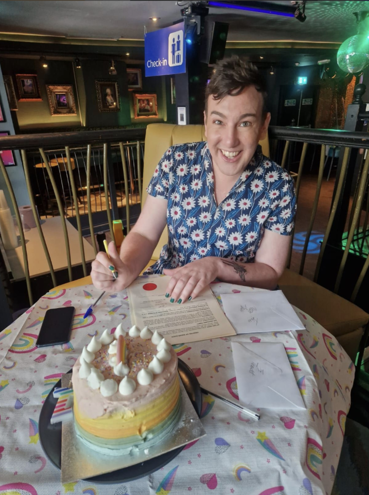 This is an image of a trans and non-binary person signing a document. There is a cake on the table. She is wearing a blue dress with white accents. 