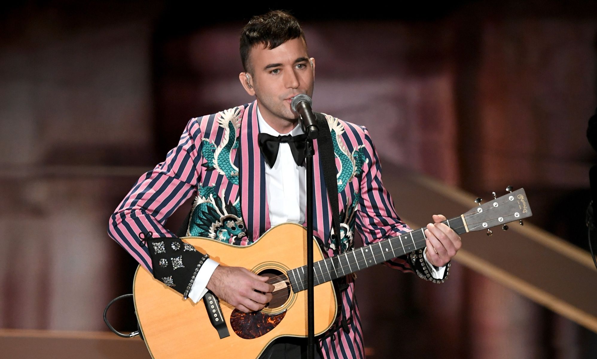 Sufjan Stevens wears a pink and black striped suit jacket as he plays the guitar and sings into a microphone on stage