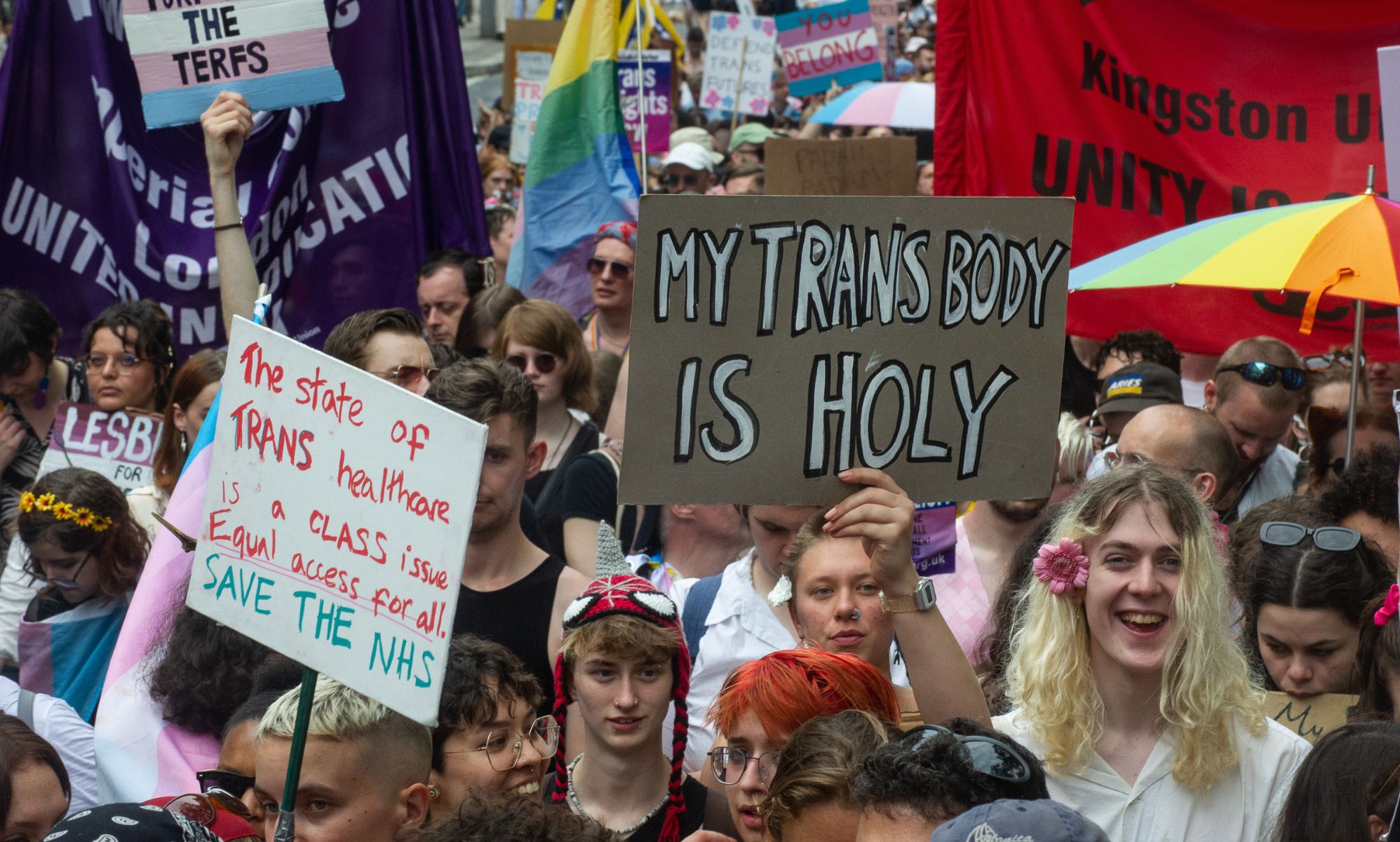 A crowd of people gather together in protest for the trans community. One person holds up a sign reading 