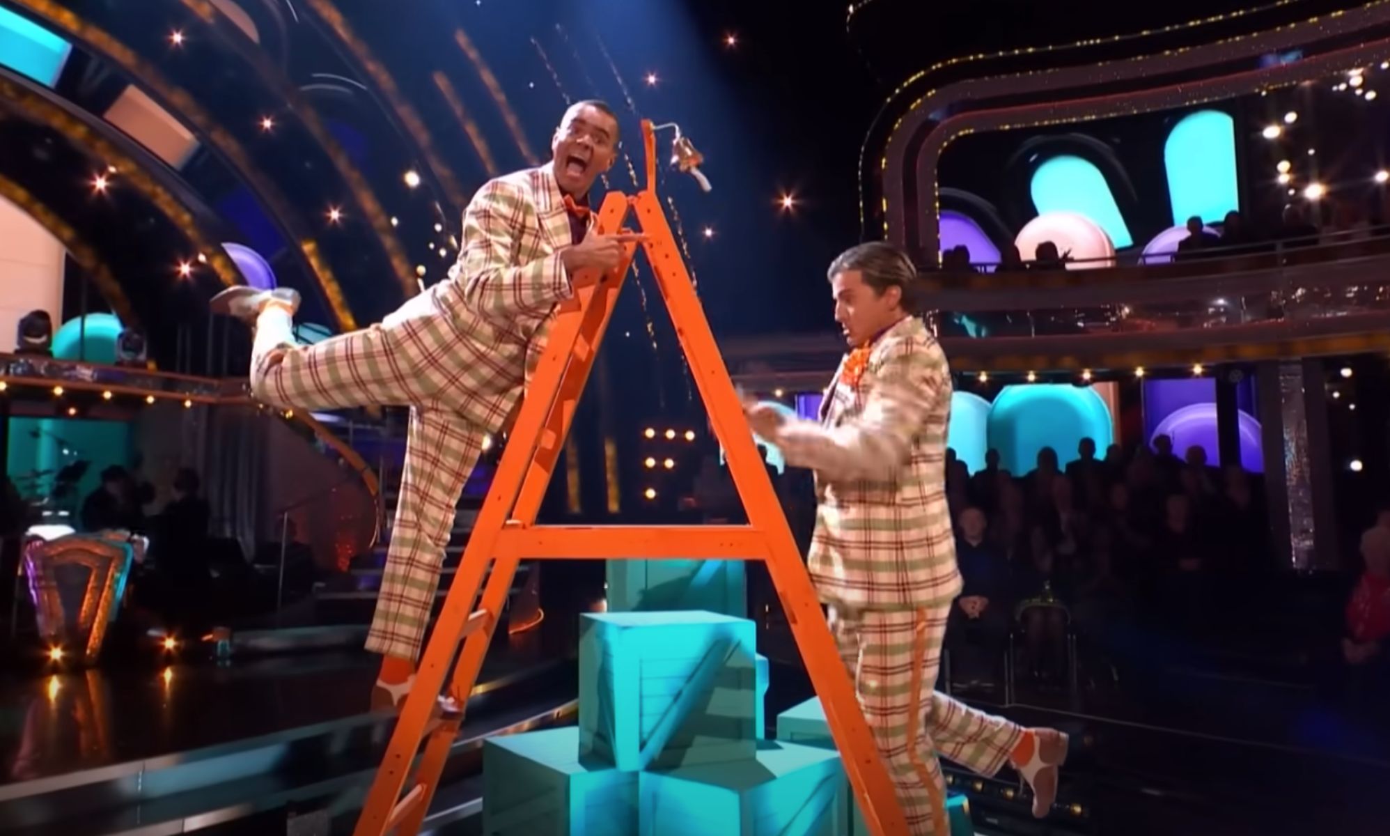 Layton Williams and Nikita Kuzmin wear matching plaid suits as they stand on a ladder during a performance on BBC