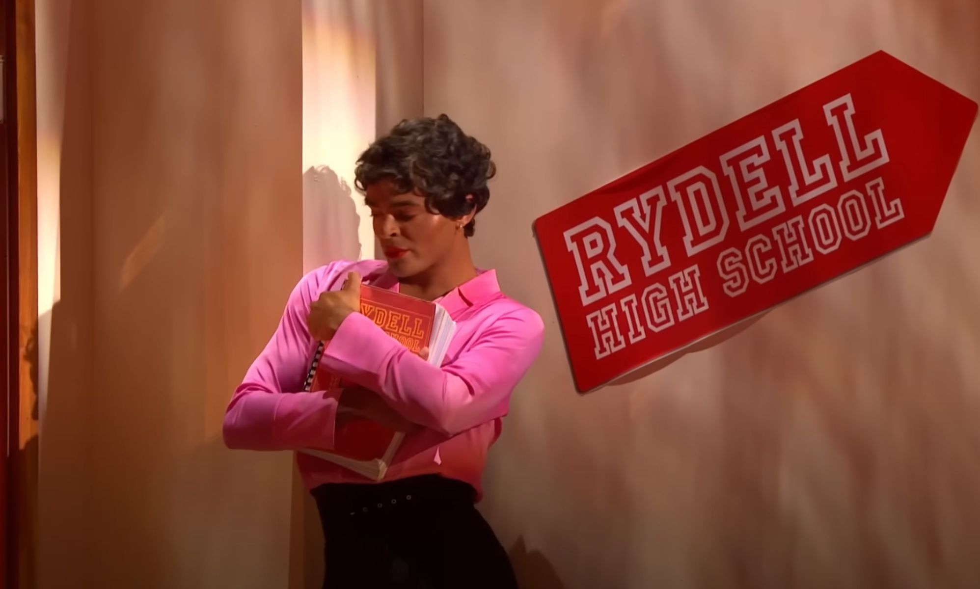 Layton Williams wears a pink shirt and black skirt as he holds a book during a performance on BBC