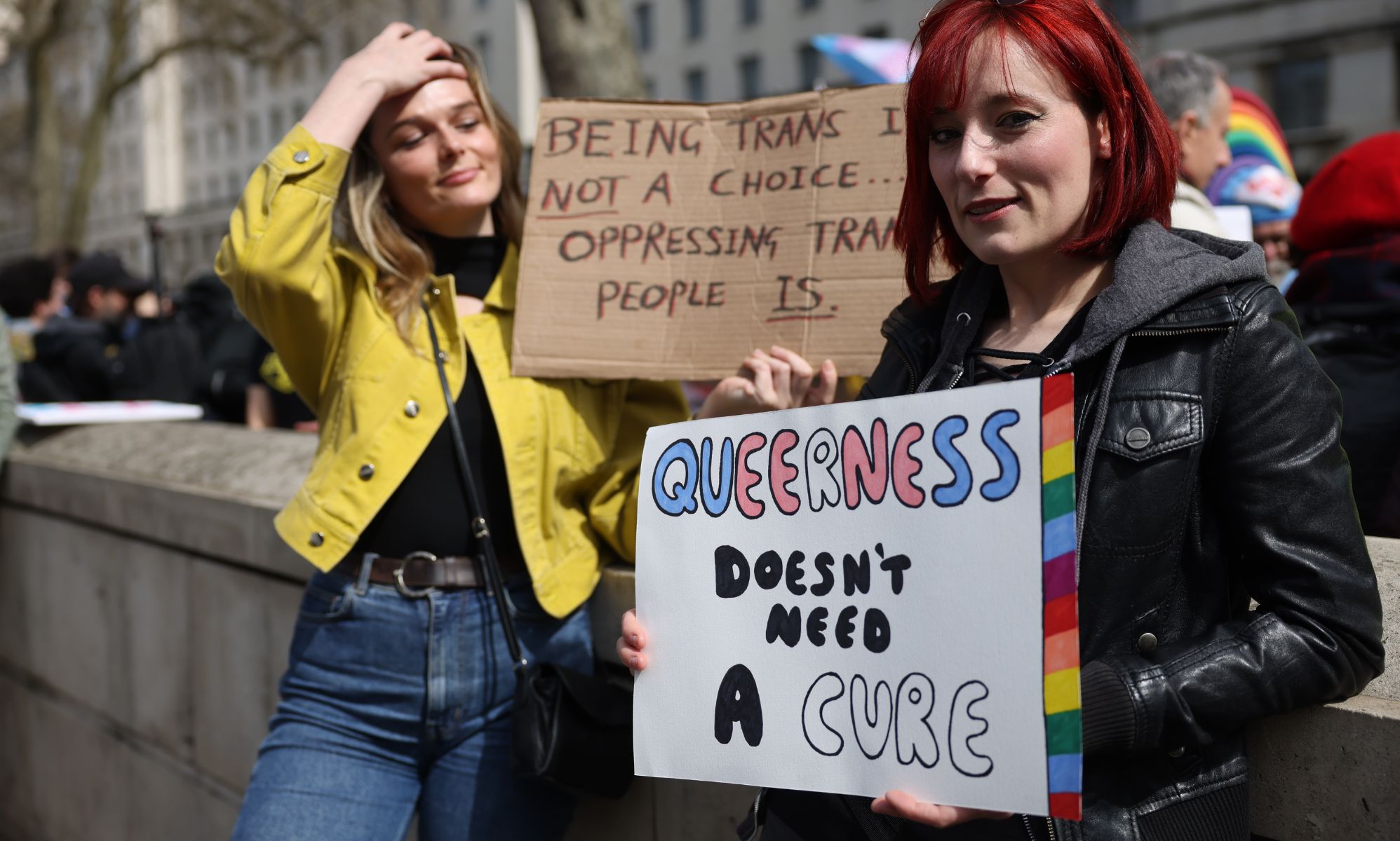 Two people stand side by side during an LGBTQ+ rights protest to ban conversion therapy. One person holds up a sign reading 