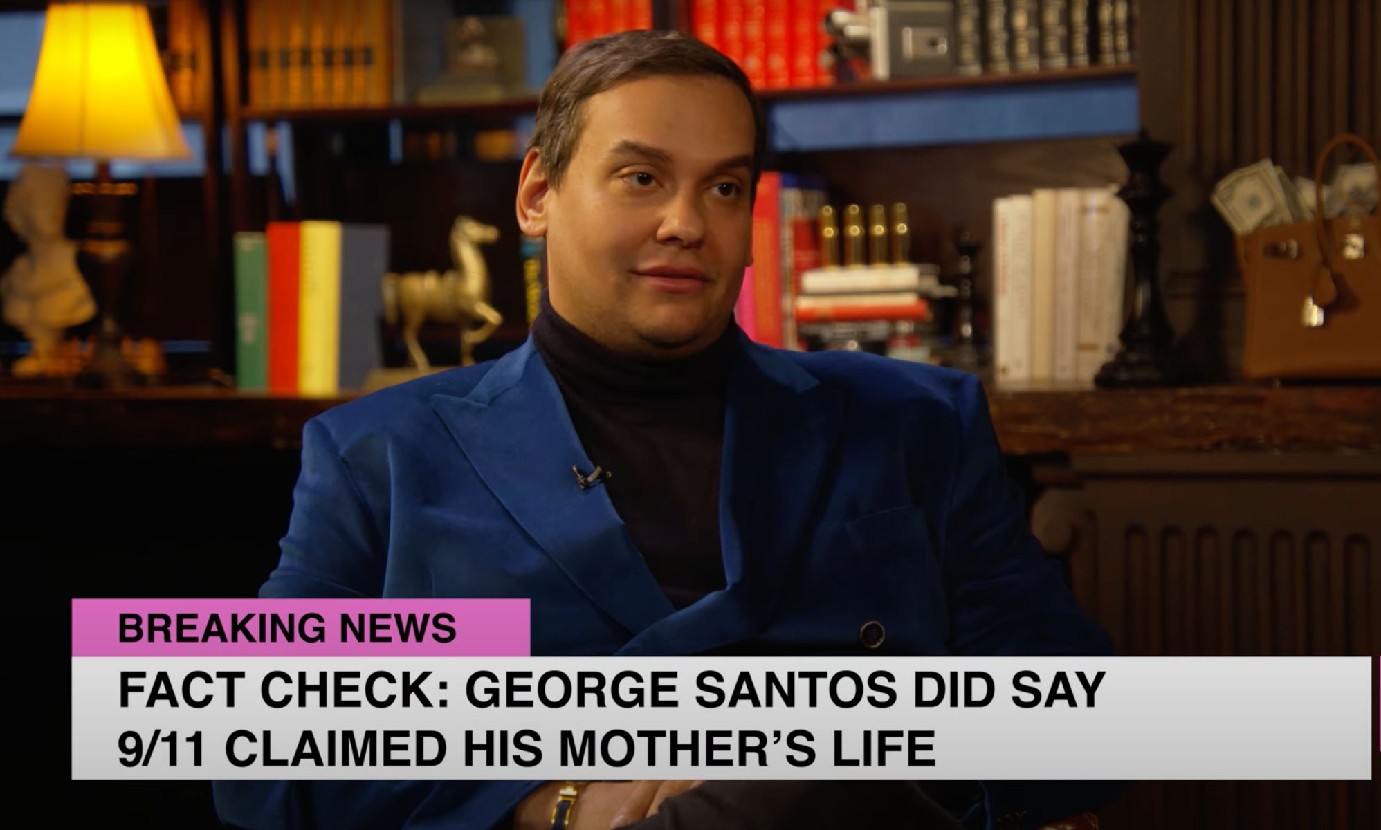 George Santos wears a black shirt and blue jacket as he sits in front of a bunch of books during an interview with Ziwe. In this frame, there