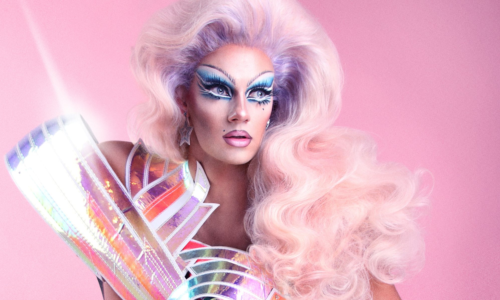 A person in drag wearing a pink wig poses infront of a pink background.