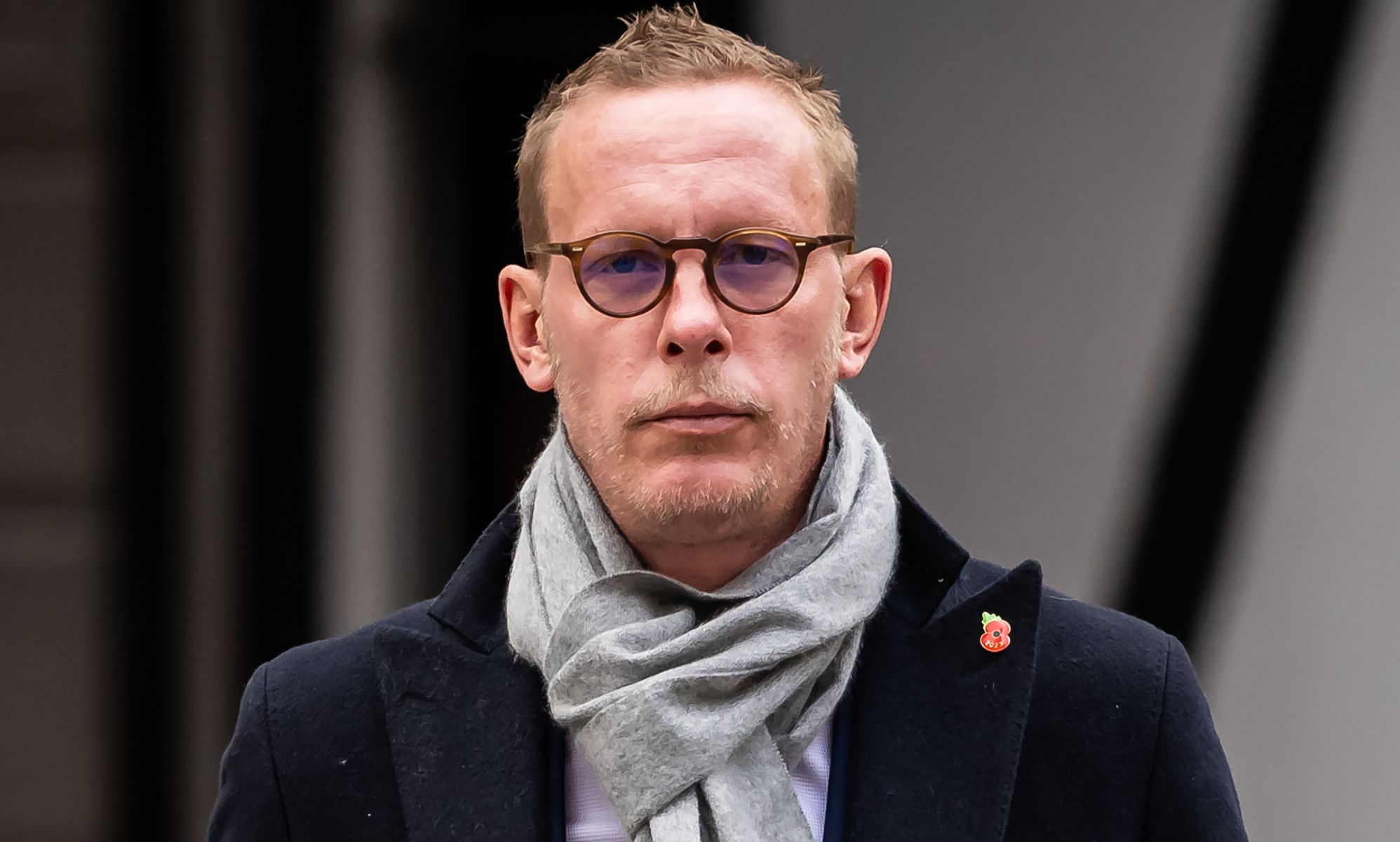A man stands wearing a scarf, a coat and some glasses.