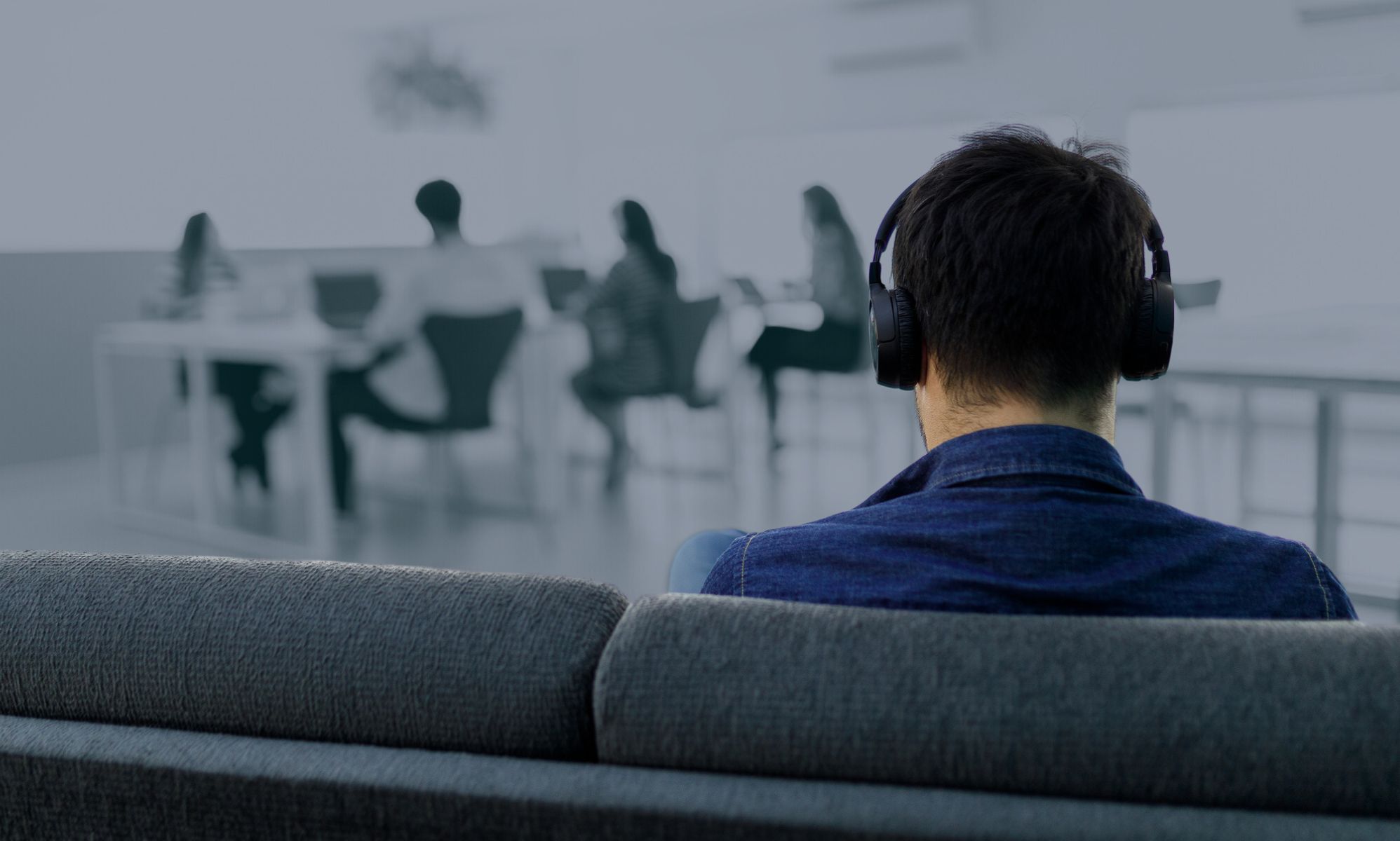 This is an image of the back of a male presenting person sitting alone on a couch and is wearing noise-cancelling headphones. In the background there are a group of colleagues seated at a table. 