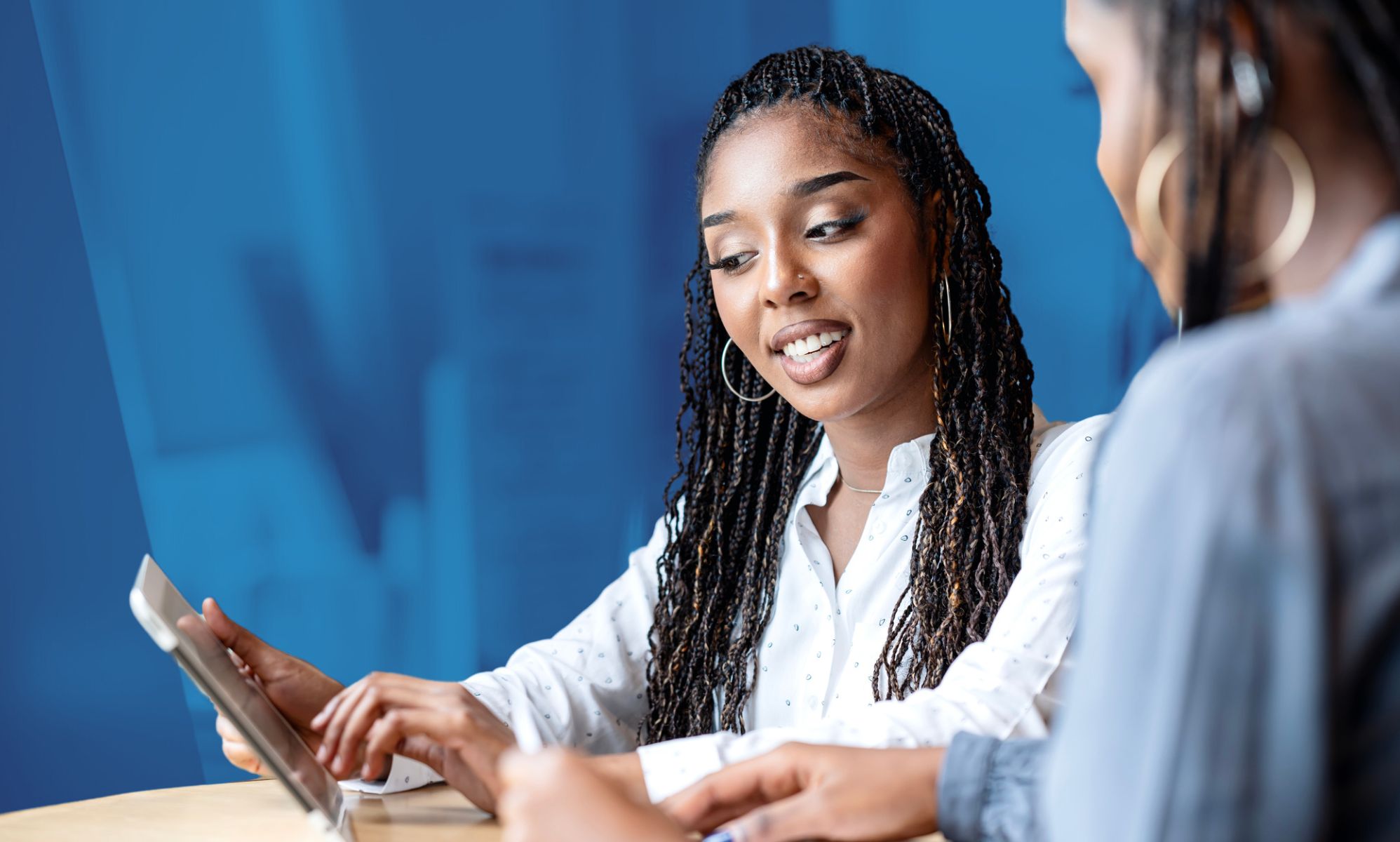 This is an image of 2 Black women at work. The are both looking down at a tablet.