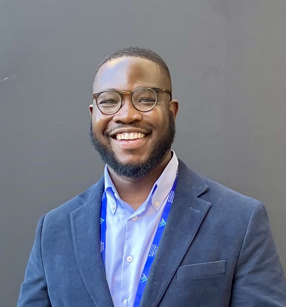 This is an image of a young Black man. He is wearing a blazer with a light blue shirt. He has round glasses on.