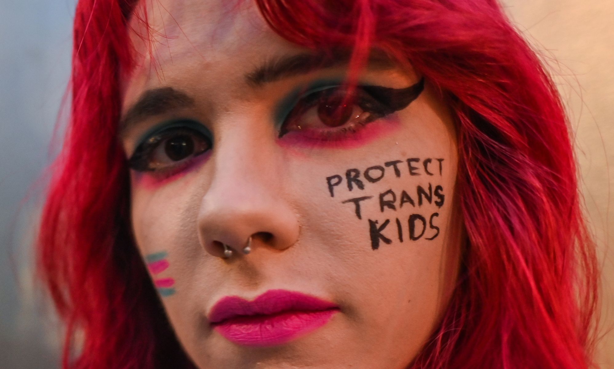 A close up of a trans activist with the words &quot;protect trans kids&quot; written on their face.