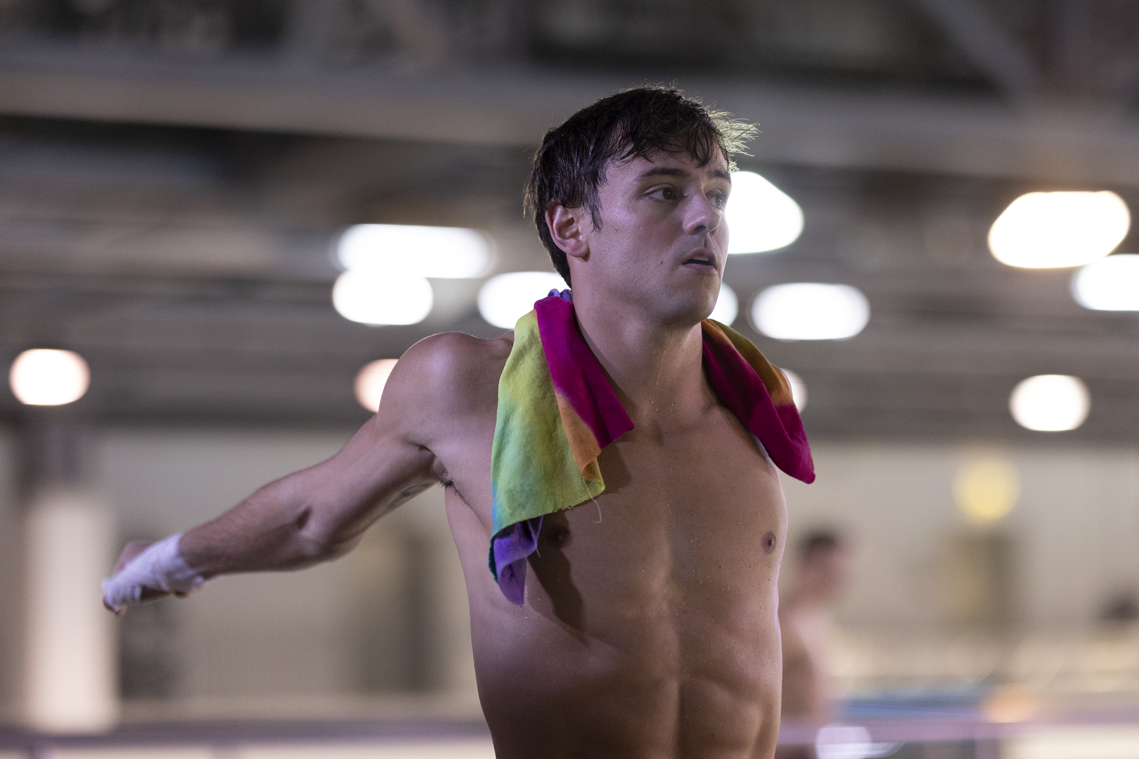 Tom Daley stretching his arms behind his back with a rainbow towel around his neck.