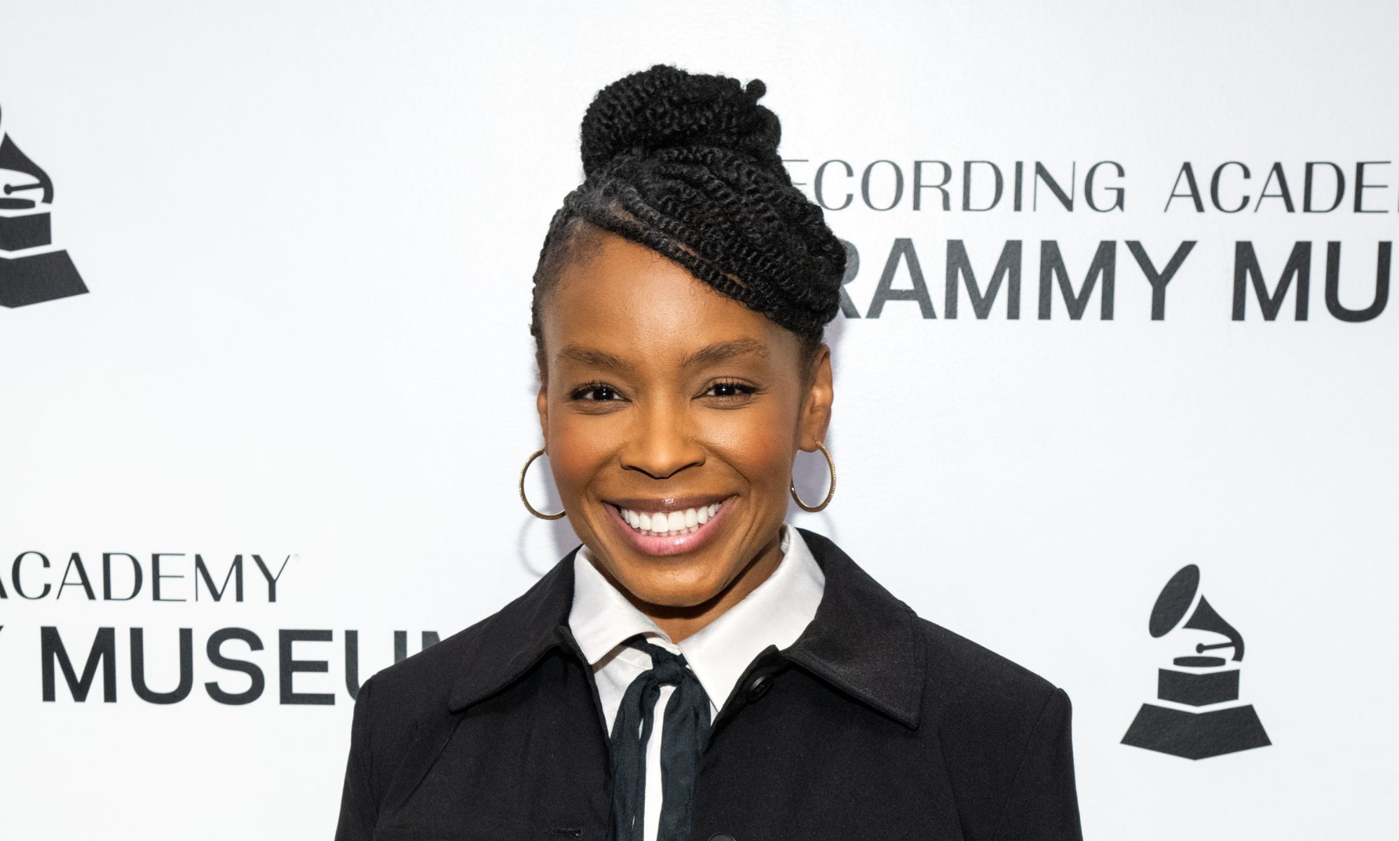 Comedian Amber Ruffin in a black suit jacket, white shirt and black tie. She is smiling while standing on the Grammy 2024 red carpet.