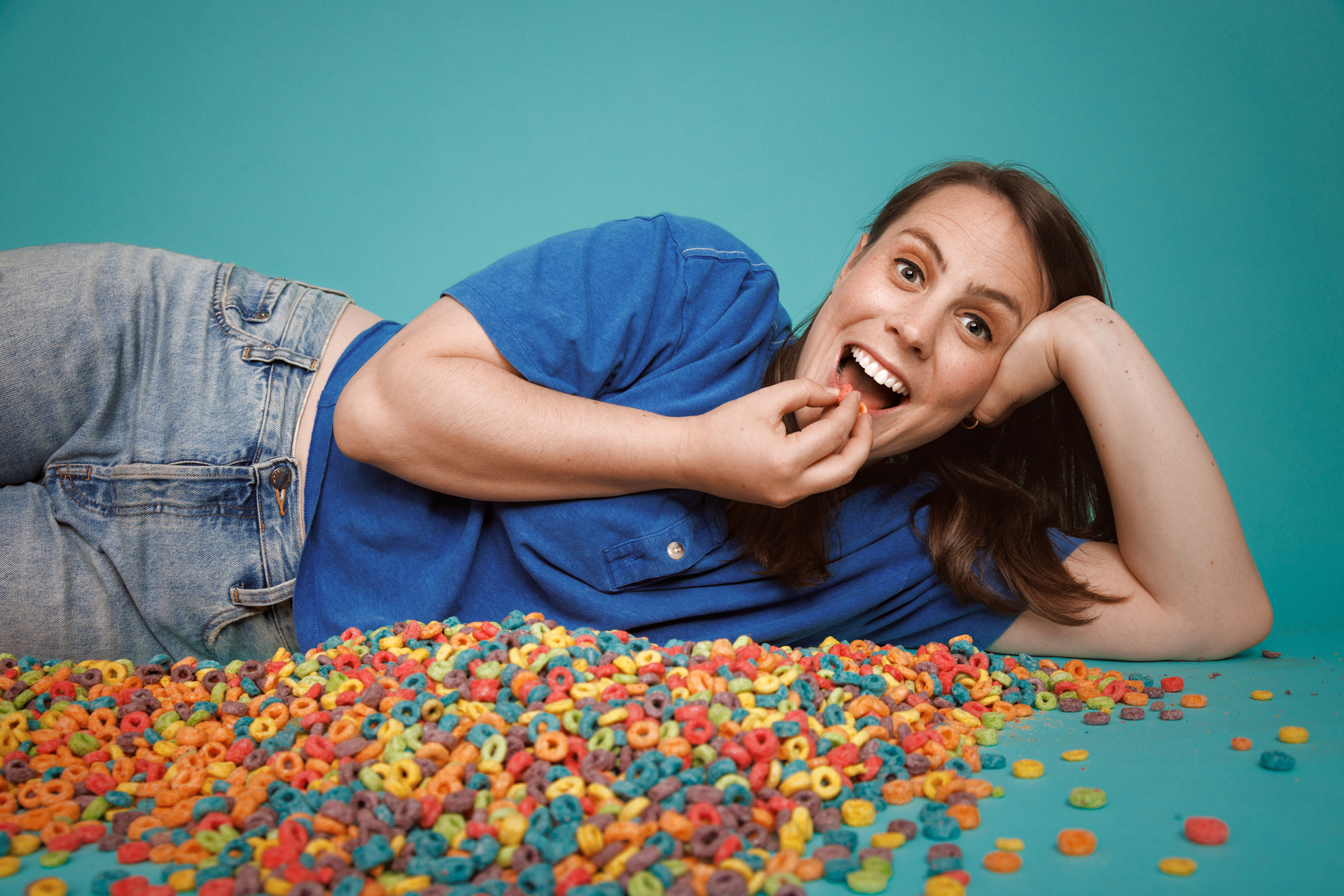 Olivia Levine: Unstuck. Olivia lyng on the ground against a light blue background with cheerios all over the floor, she
