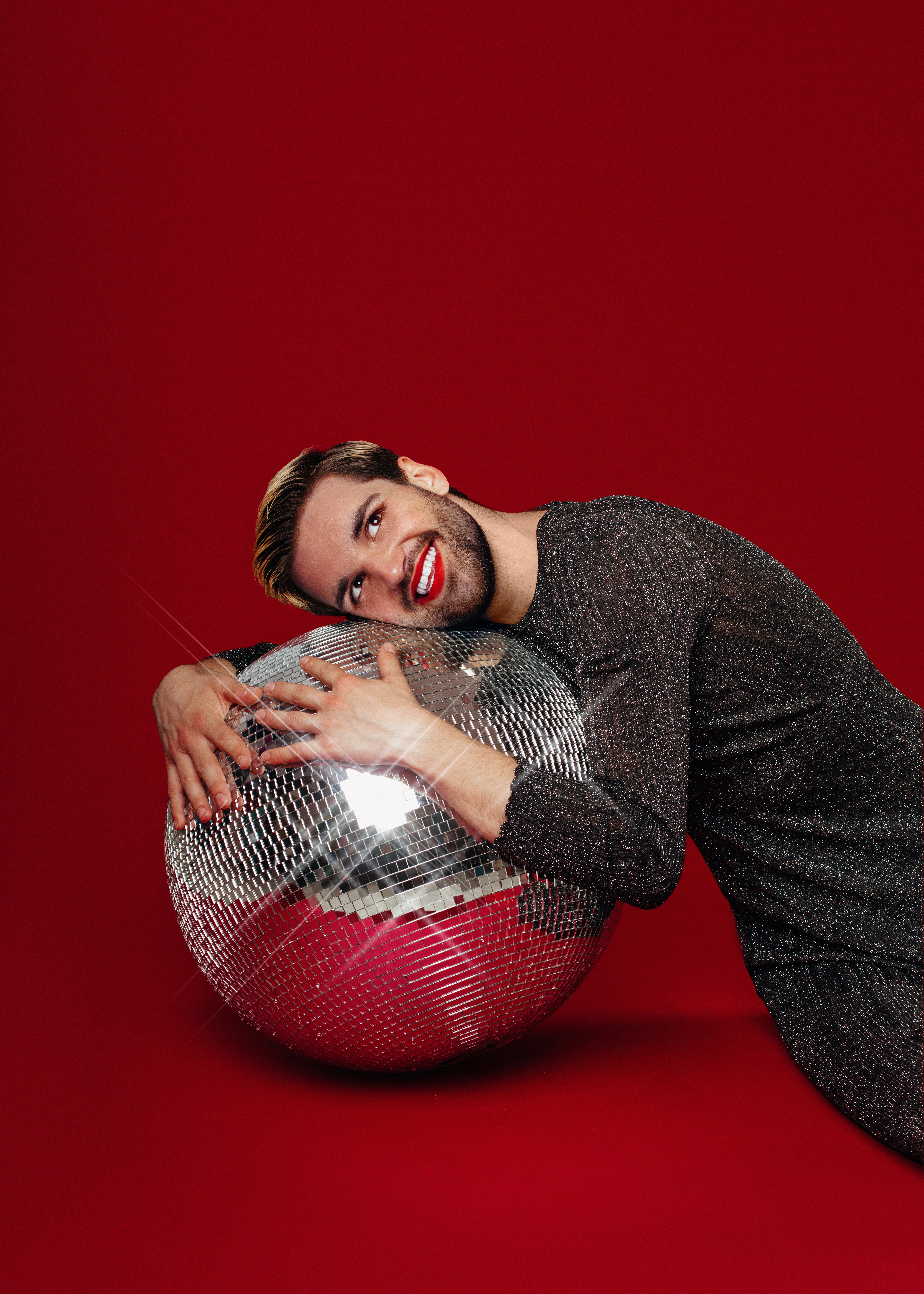 Will Owen in a sparkly look with red lipstick hugging a mirrorball.