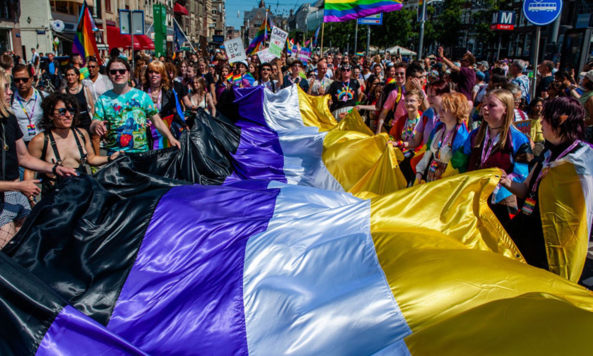 People are holding a large non-binary flag during a Pride march in Amsterdam. From left to the right the colours are black, purple, white and yellow.