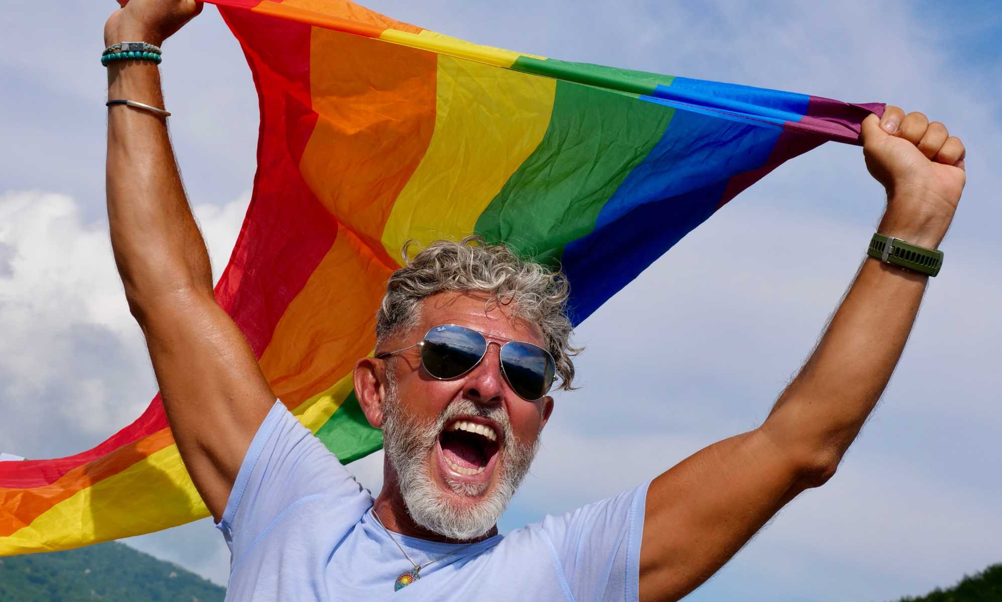 Portrait of a gray-haired elderly Caucasian man with a beard and sunglasses holding a rainbow LGBTQIA flag against a sky background, shouts in protest, Celebrates Pride Month Coming Out Day