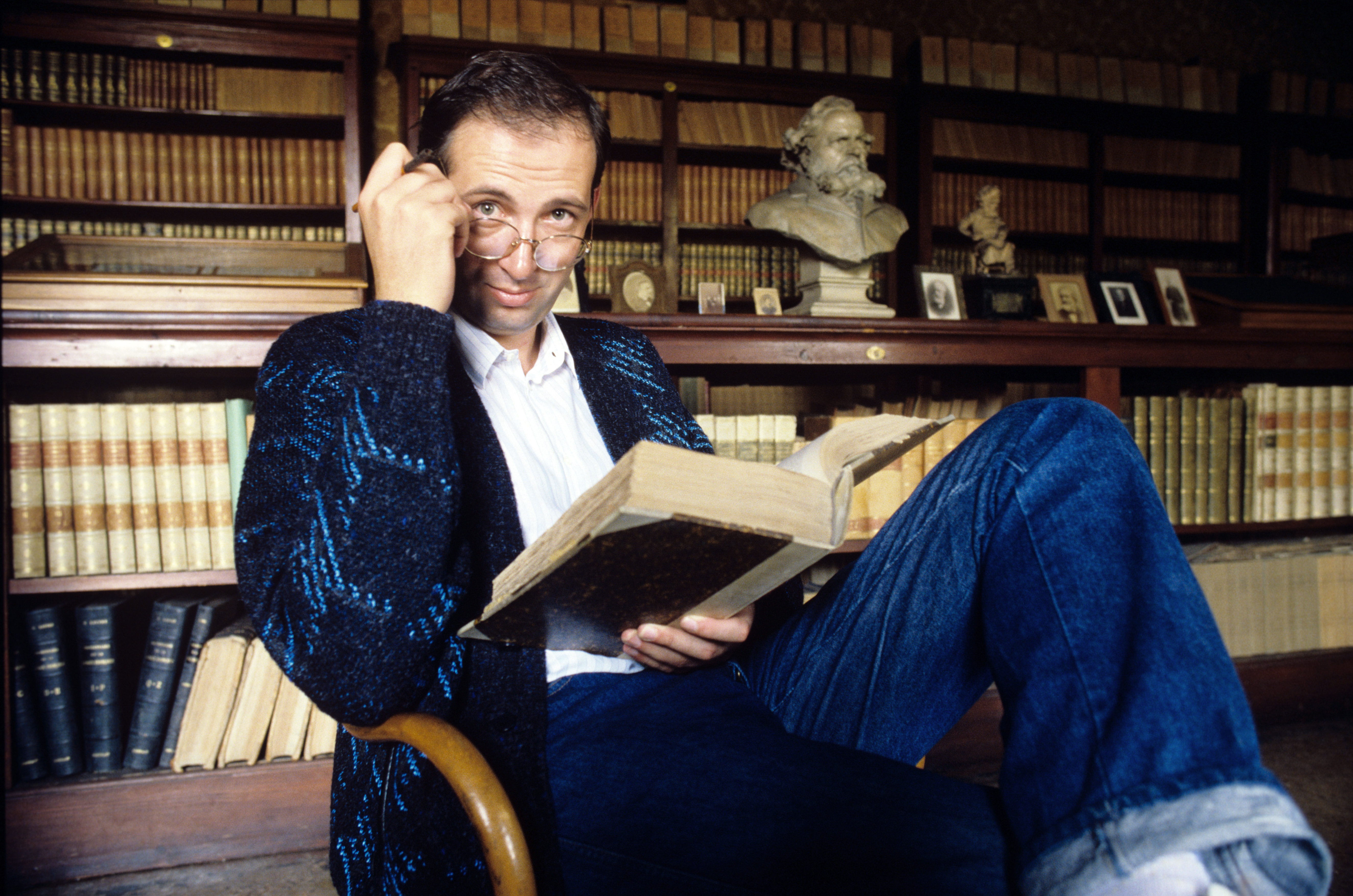 Italian writer Pier Vittorio Tondelli adjusts his glasses while reading a book, sitting on an armchair; behind him a shelf of his furnished library