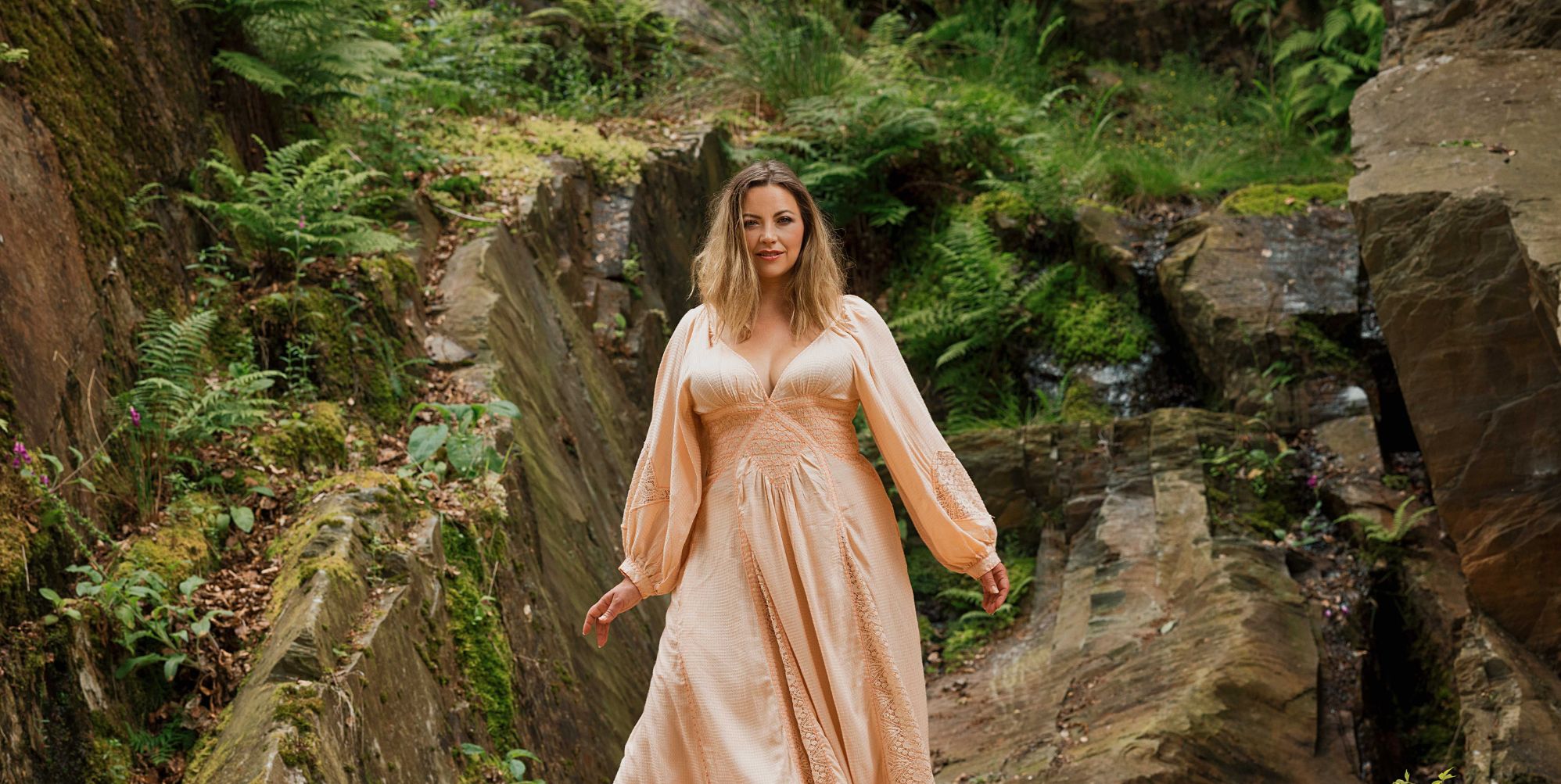 Charlotte Church in a peach dress standing against rocks and folliage.