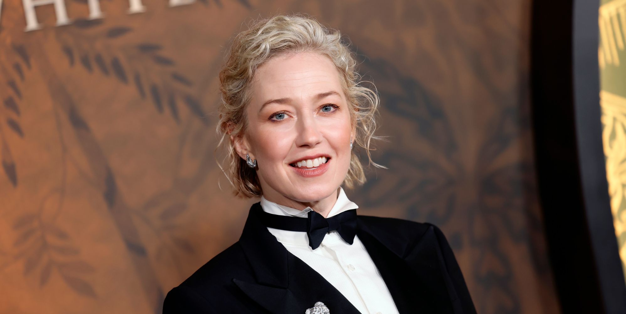 Carrie Coon at the White Lotus premiere in a suit and bow tie against a brown backdrop.