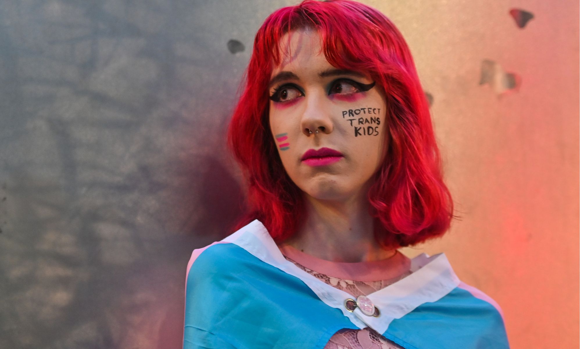 A person with red hair leaning against a wall with face paint which reads &quot;protect trans kids.&quot;