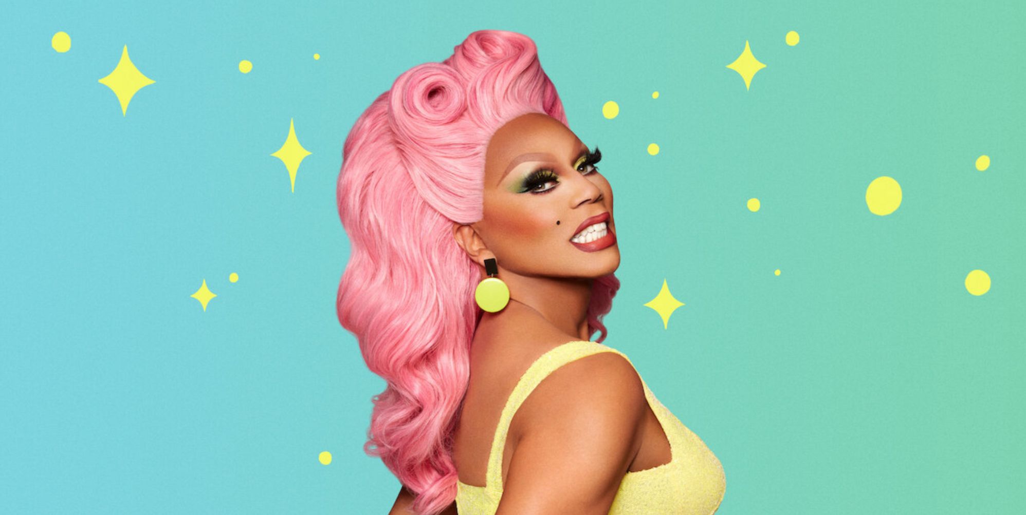 RuPaul smiling while wearing a pink wig and yellow top, stood against a mint background.