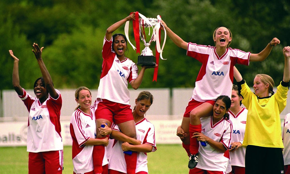 Jess (Parminder Nagra) and Jules (Keira Knightley) in red and white football kits both holding a trophy while being on the shoulders of their teammates.