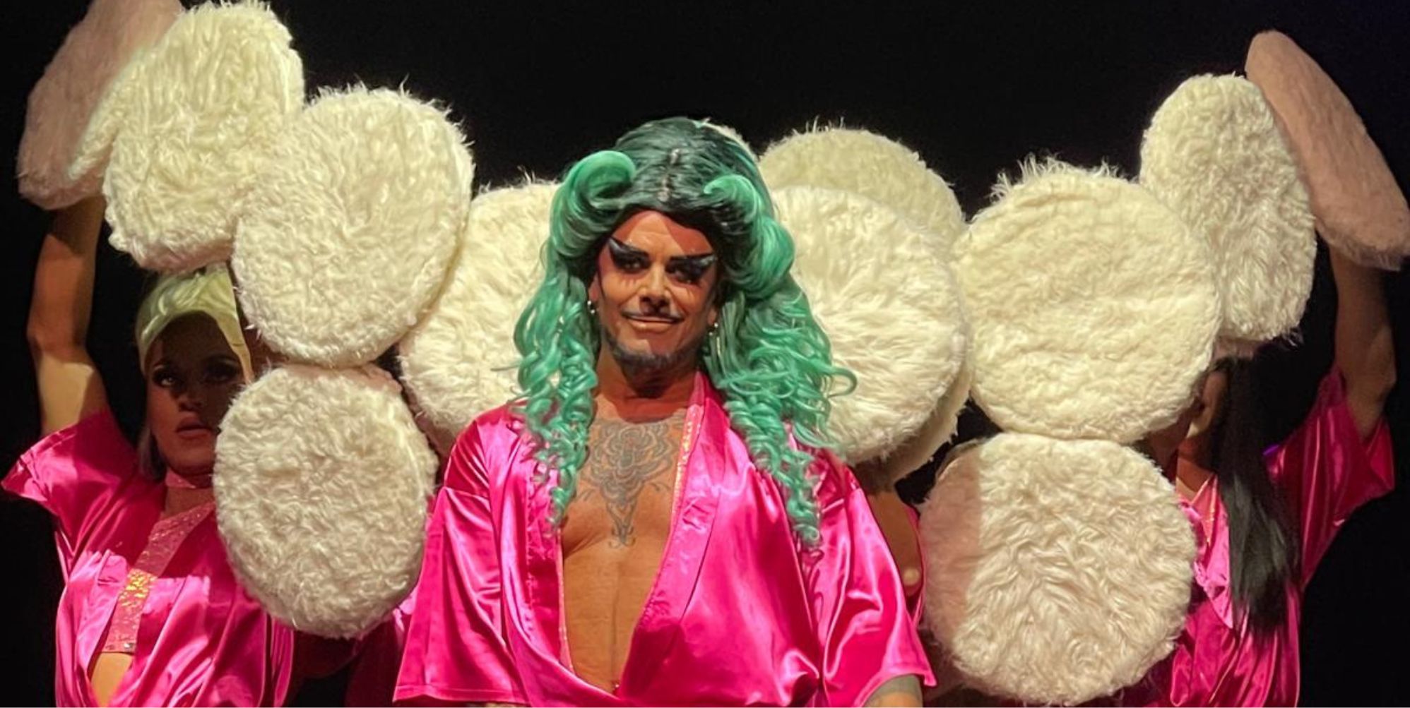 A drag performer in a pink robe and green wig surrounded by large powder puffs.