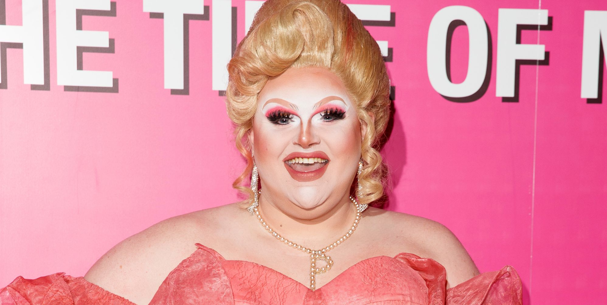 Pixie Polite in a pink dress and blonde wig posing and smiling against a pink and white background.