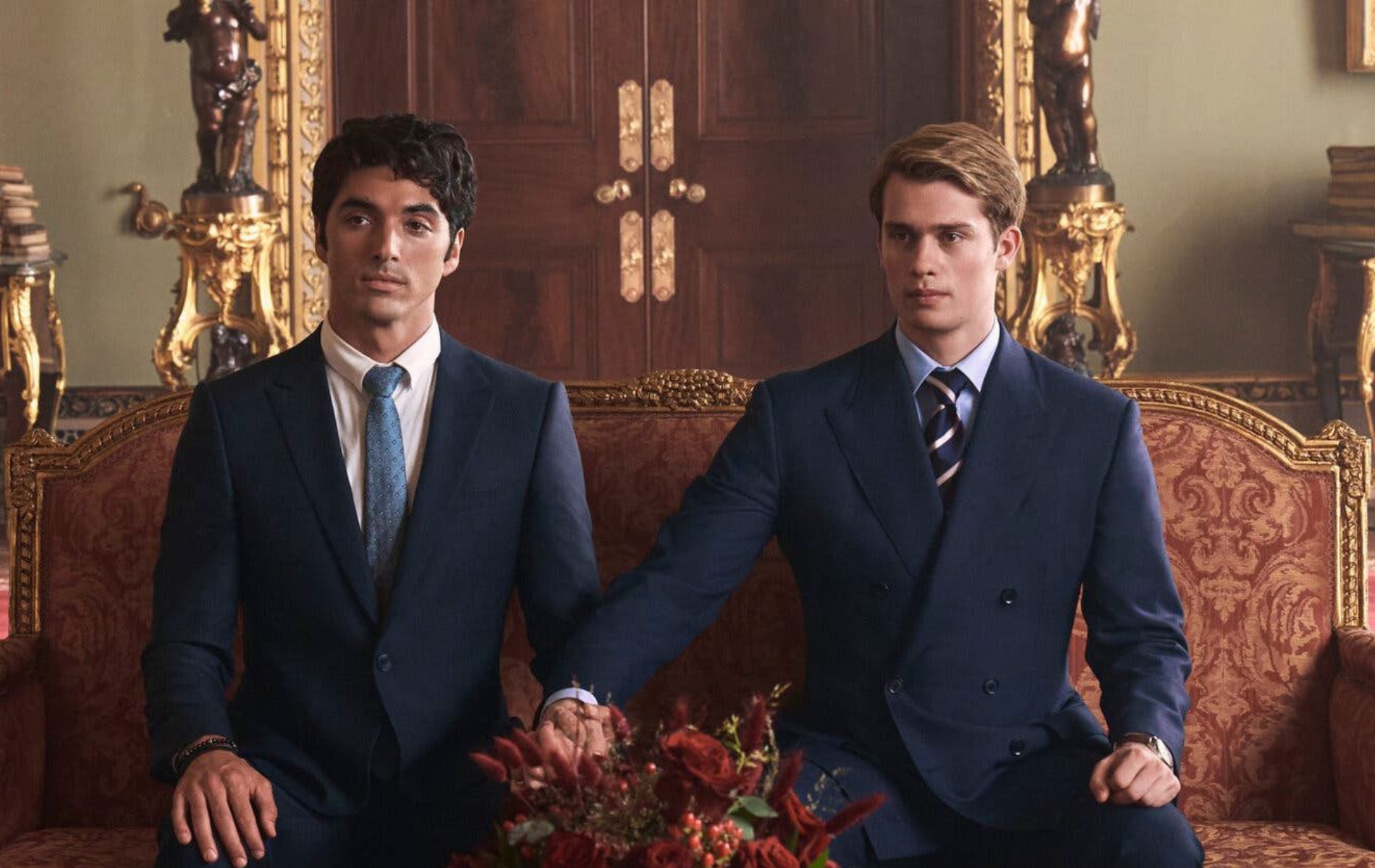 Red, White &amp; Royal Blue still: Alex (Perez) and Prince Henry (Galitzine) sat on a royal red and gold sofa, both wearing suits. Henry has stretched his arm to hold Alex