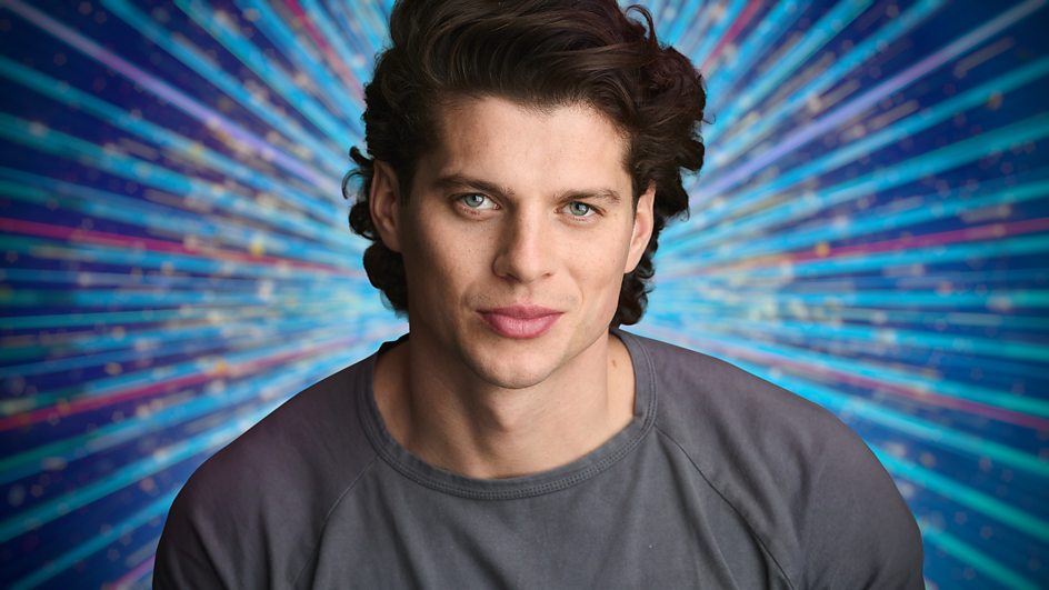 Lewis Cope wearing a grey t shirt in front of a blue streamer Strictly background