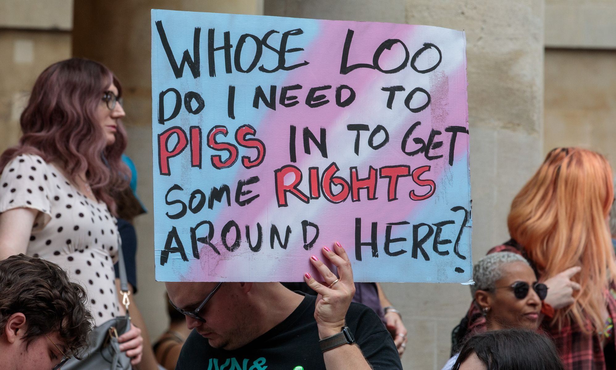 A person holding a sign during a trans protest in the UK, which reads &quot;Whose loo do I need to piss in to get some rights around here?&quot;