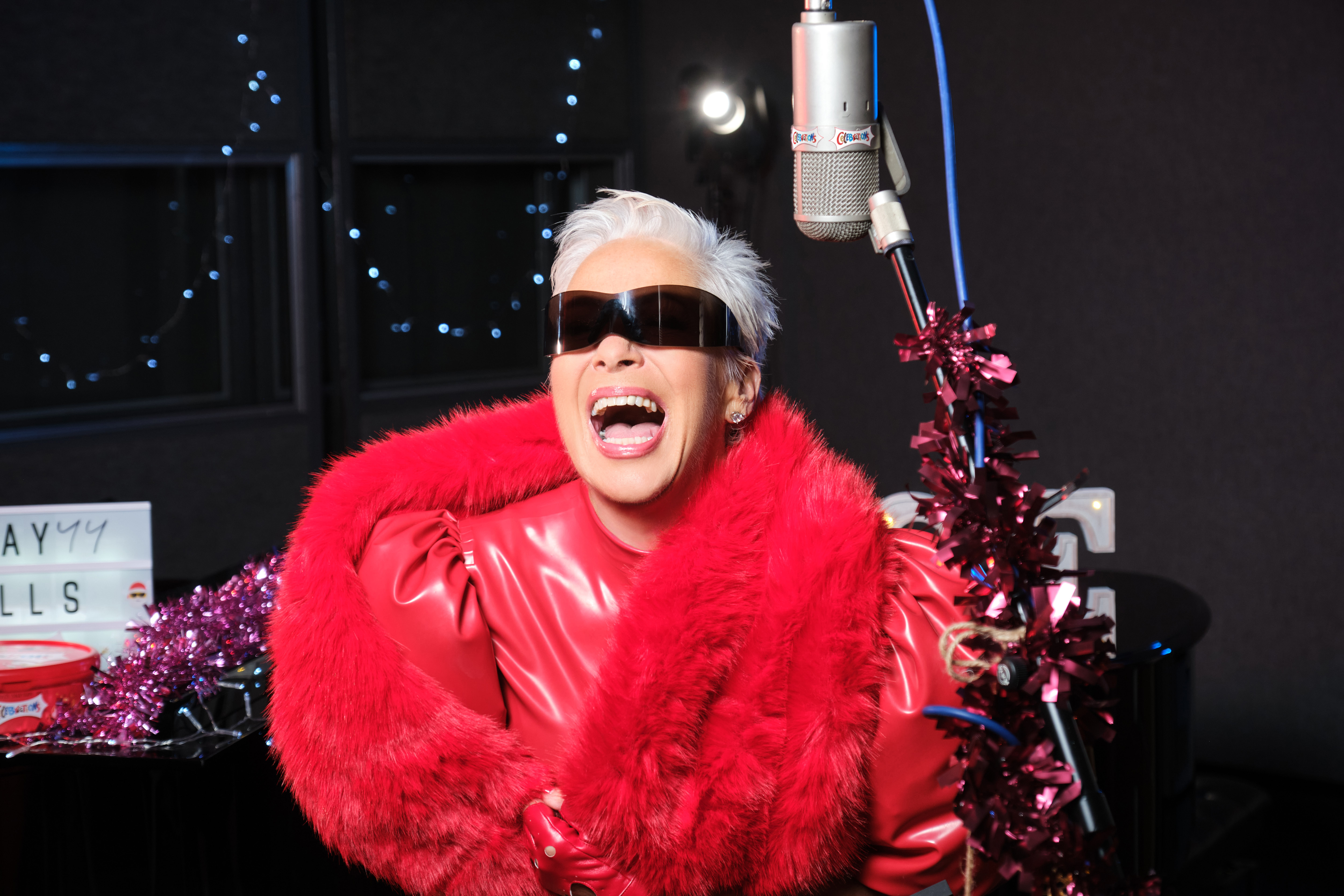 Denise Welch in all red and sunglasses, laughing close to a microphone.