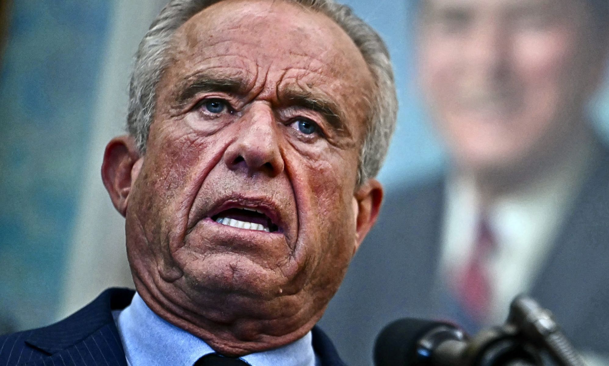 A  close-up of a 71-year-old man in a suit and tie speaking inside a government building.