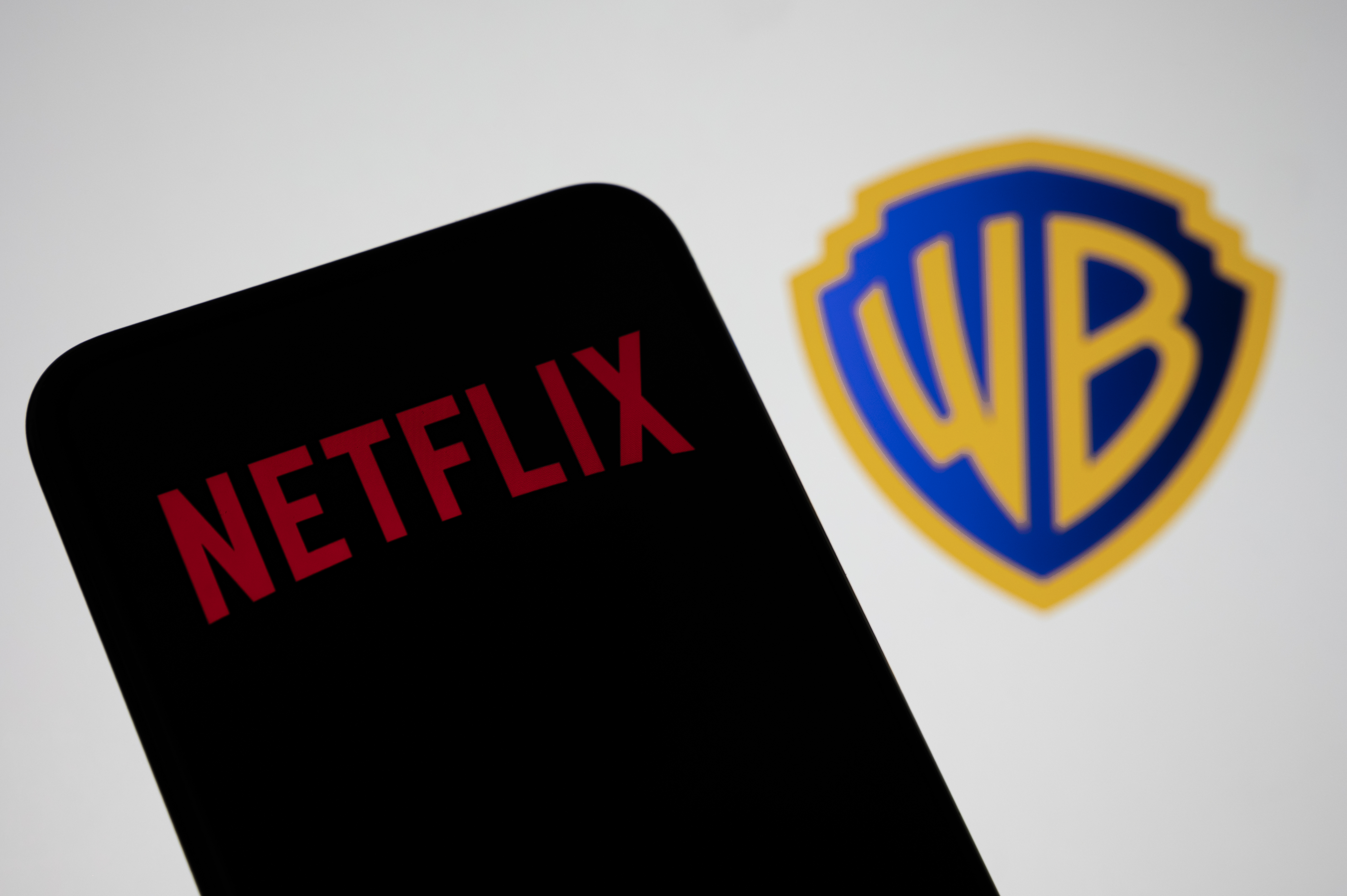 In this photo illustration, the Netflix logo appears on a cell phone with the Warner Bros. logo in the background, in Creteil, France, on December 10, 2025. (Samuel Boivin/NurPhoto via Getty Images)