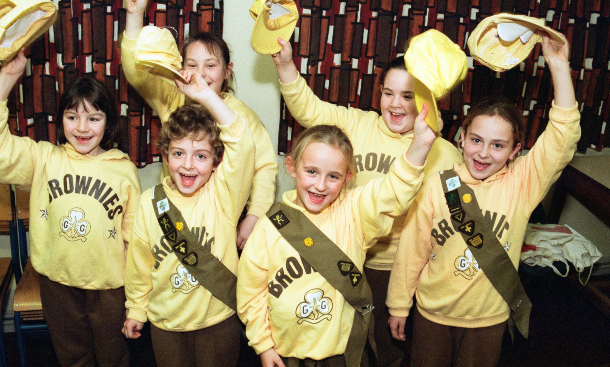 A group of children in yellow and brown Brownies uniforms holding up their hats for a picture.