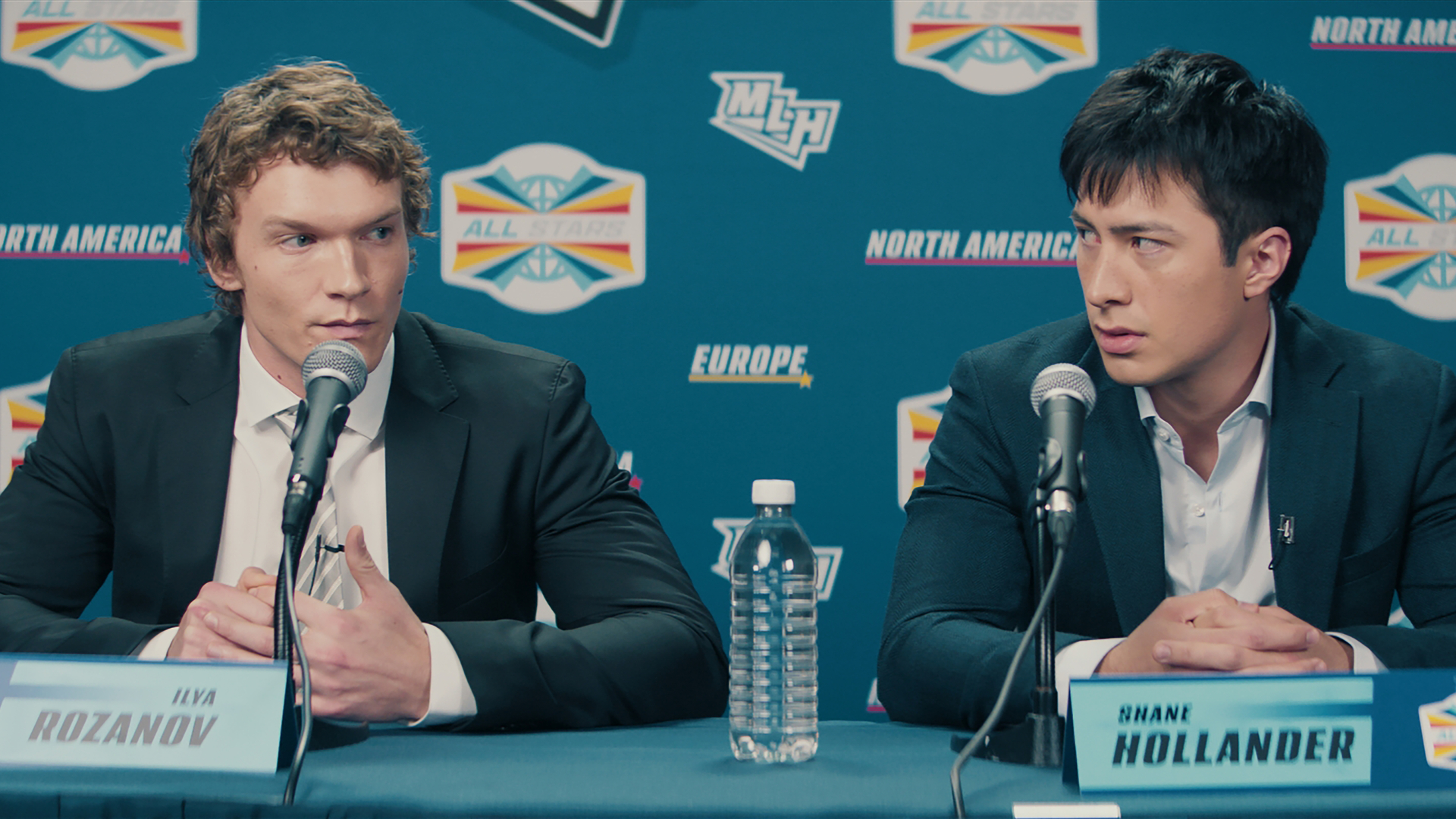 Heated Rivalry (L to R) - Connor Storrie as Ilya Rozanov and Hudson Williams as Shane Hollander. Sitting at a press conference in suits